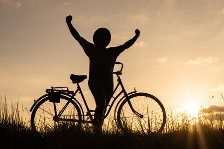 Silhouette of a happy cyclist man in a hat with his arms raised up, a symbol of freedom, standing in a meadow with a bicycle during sunset. Healthy active lifestyle concept.の写真素材