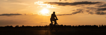 Long Banner. Close-up silhouette of a bicycle with cyclist's legs at sunset. Background of a field in the countryside at dawnの写真素材