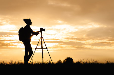 Silhouette of a female photographer with a backpack and a professional photo-video camera on a tripod, standing in a meadow in a field in the countryside. Background of sunset on sky with clouds.の写真素材