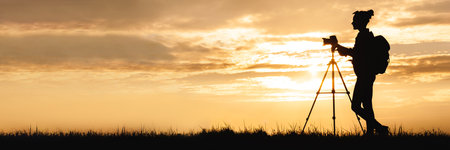 Long banner silhouette of a female photographer with a backpack and a professional photo-video camera on a tripod, standing in a meadow in a field in the countryside. Background of sunset on sky with clouds.の写真素材