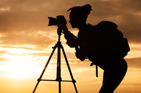 Silhouette of a young woman photographer with a backpack and a professional photo camera or camcorder on a tripod. Background of sunset sun on the sky with clouds.の写真素材