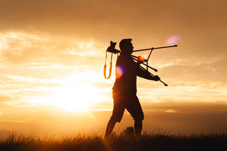 Silhouette of a man walking through a grassy field at sunset with a camera and tripod over his shoulder. The scene conveys freedom, exploration, and a passion for photography in natural light.の写真素材