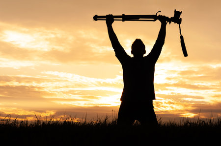 A silhouette of a man triumphantly raising a camera and tripod above his head during a golden sunset. The image conveys themes of achievement, freedom, creative success, and passion for visual storytelling.の写真素材