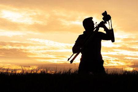Silhouette of a man standing in a grassy field, holding a tripod with a camera over his shoulder during sunset. The image reflects themes of adventure, creativity, photography, and the peaceful beauty of nature.の写真素材