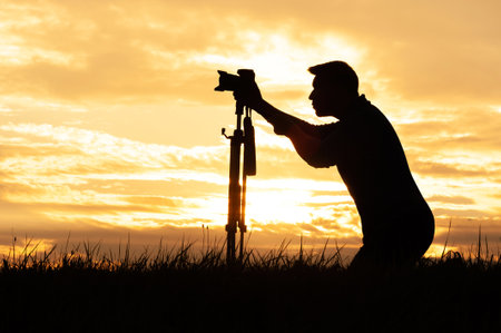 A silhouette of a man kneeling and adjusting a camera on a tripod against a vibrant sunset sky. This...