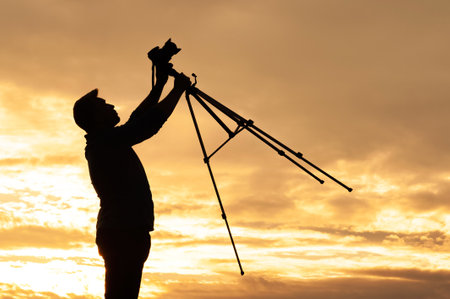 Silhouette of a man raising a camera mounted on a tripod into the air at sunset. This dramatic moment highlights outdoor photography passion and the beauty of nature at golden hour.の写真素材