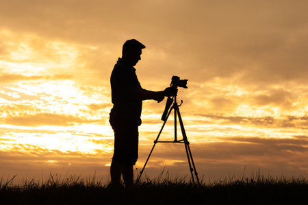 Silhouette of a photographer with a professional camera standing on a tripod. Against the background of the sunset skyの写真素材