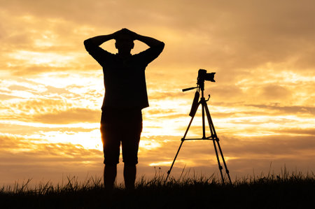 Silhouette of a man standing with hands on his head next to a tripod-mounted camera at sunset. The...