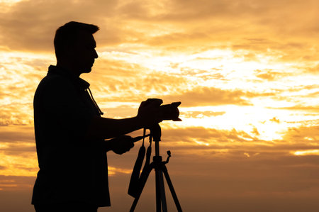 A silhouette of a male photographer adjusting his camera mounted on a tripod during a vibrant...