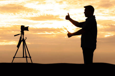 A male photographer in silhouette frames a shot with his thumbs in front of a camera on a tripod during sunset. The scene emphasizes creativity, composition, and outdoor photography during golden hour.の写真素材