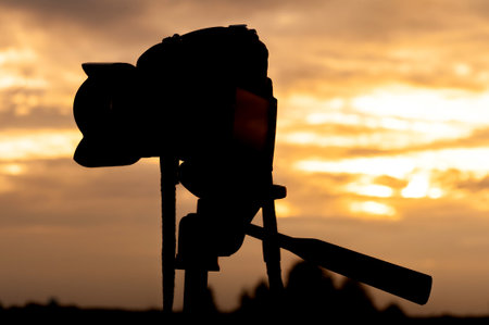 A DSLR camera on a tripod is silhouetted against a vivid sunset sky with warm golden tones. The image captures a peaceful outdoor moment, ideal for concepts of photography, equipment, and nature.の写真素材