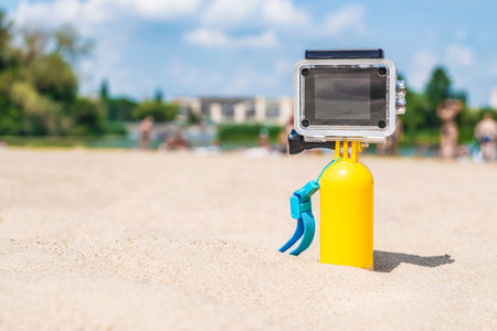 Close-up Mock-up of a waterproof action camera with a yellow float standing on a sandy beach. Background in bokeh people green trees blue sky and cloudsの写真素材