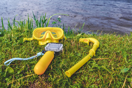 Close-up action camera with waterproof body and yellow float with underwater mask and diving snorkel. Shore background with water and green grassの写真素材