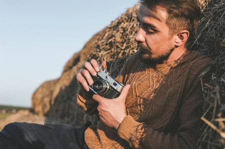 Man setting up retro photo camera outdoors while sitting on big bales of hayの写真素材
