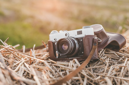 A retro camera with a brown case lies on a haystack. Background of a field with green grass, at sunset with a sunbeamの写真素材