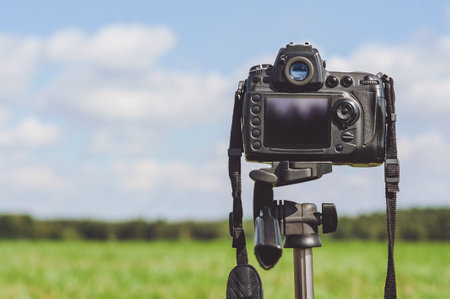 Close-up of a professional photo video camera on a tripod stands in a field. Background of forest and skyの写真素材