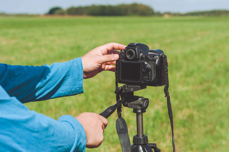 Close-up of a professional photo-video camera in the hands of a man. Against the backdrop of a green landscape with treesの写真素材