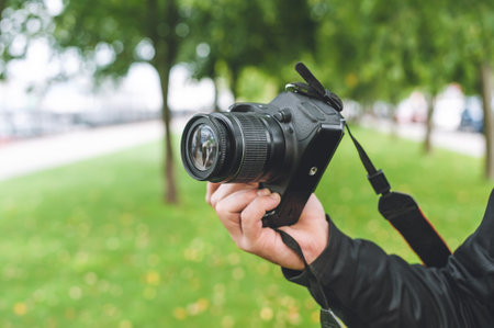 Guy man holds a professional photo-video camera in his hand. Background of a beautiful park with a green lawn and treesの写真素材