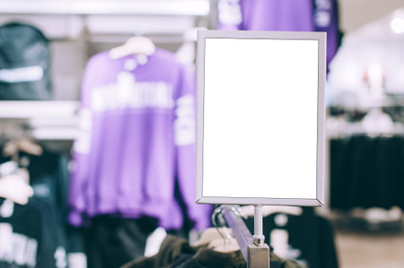 Blank mockup white rectangular sign with a silver frame in the foreground of a clothing store, with a blurred purple long-sleeved shirt hanging in the background. This provides space for advertising or information.の写真素材