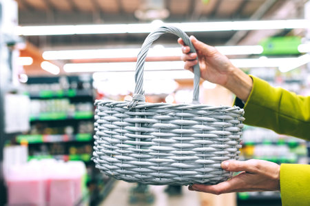 Person's hands holding a light gray oval wicker shopping basket in a retail store aisle with blurred shelves and lighting in the background. The focus is on the basket being held.の写真素材