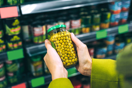 First-person view of a person's hands holding a clear glass jar filled with green peas in a supermarket aisle with blurred shelves stocked with canned goods. The focus is on selecting the peas.の写真素材