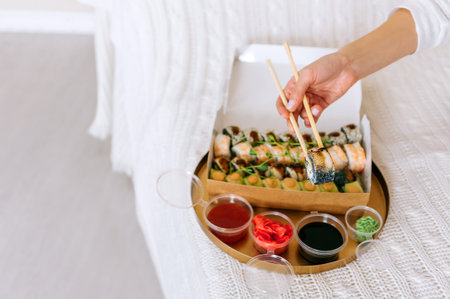 Woman holding sushi with chopsticks. Against the backdrop of a set of takeaway rolls. in a paper package on a tray with ginger and wasabi sauces in the house on the bed.の写真素材