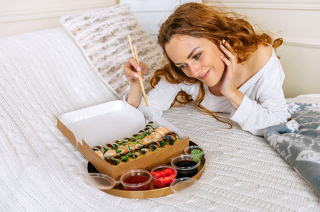A young beautiful woman eating a sushi set of rolls in a takeaway box while sitting on a bed in a bright house. Asian cuisine delicious food holds chopsticks in hand indoors red haired girl.の写真素材