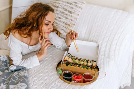 A young beautiful woman eating a sushi set of rolls in a takeaway box while sitting on a bed in a bright house. Asian cuisine delicious food holds chopsticks in hand indoors red haired girl.の写真素材