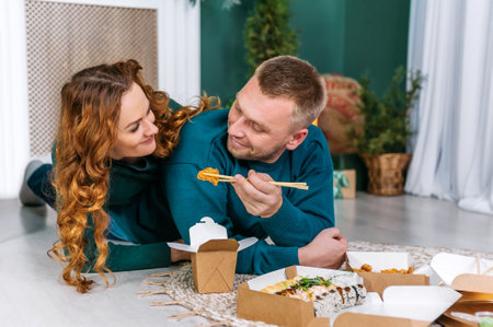 A man and a woman eat breaded shrimp nuggets and sushi from a paper take-out box while lying on the floor in the house. fast food home delivery concept.の写真素材