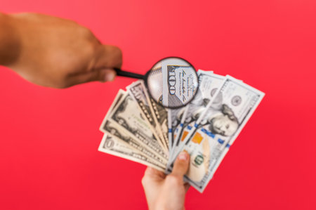 A magnifying glass in a man's hand in front of a woman's money in US dollars. On a red backgroundの写真素材