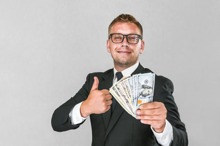 Cheerful businessman in a suit holds American dollars in his hands and thumbs up on a gray and white backgroundの写真素材