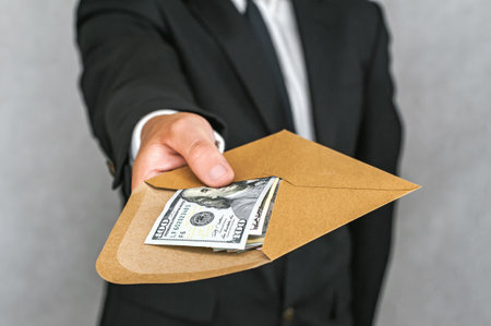 A businessman in a suit is handing over a brown envelope filled with US hundred-dollar bills. The image represents a financial transaction or a confidential cash payment.の写真素材