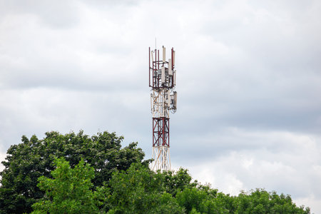 A tall cell tower with antennas stands against a cloudy sky, surrounded by lush green trees. Ideal...