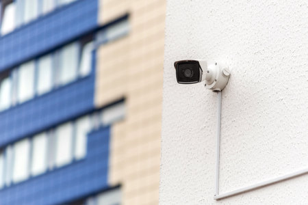 An urban security camera mounted on a white wall with a modern city building in the background. Its...