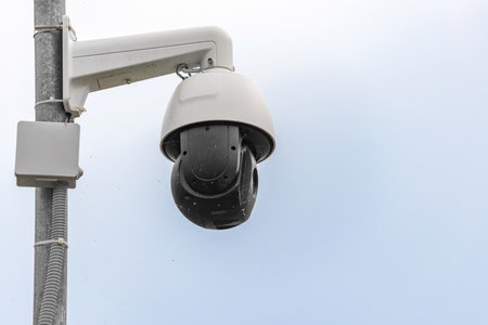 A dusty security camera covered in spiderwebs, mounted on a pole against a cloudy sky. The neglected...