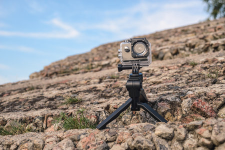 An action camera in a protective case mounted on a mini tripod stands on a rocky surface with a...