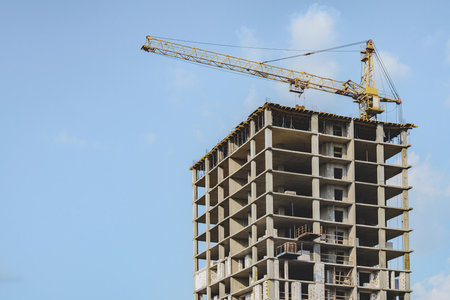 A yellow crane atop a high-rise building under construction, set against a clear blue sky with scattered clouds. Perfect for urban development and architecture themes.の写真素材