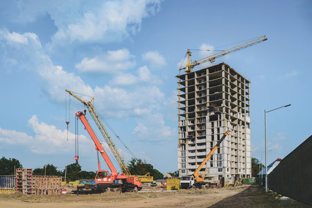 A bustling construction site features cranes and a high-rise building under construction. Blue sky with clouds enhances the urban development scene.の写真素材