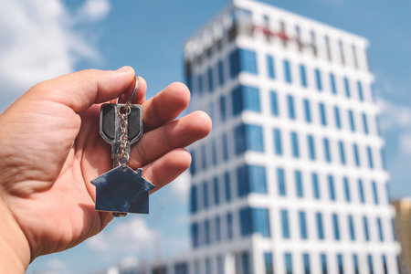 A hand holds new home keys with a house-shaped keychain against a modern building backdrop. Ideal for real estate and property investment content.の写真素材