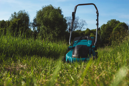 A vibrant turquoise electric lawn mower is positioned in a lush field of tall green grass,...