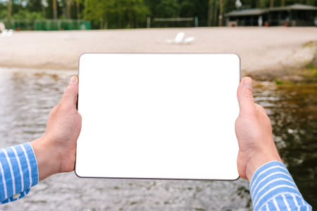 A person in a striped shirt holds a blank tablet by a lake with a sandy beach and greenery. Ideal for tech and outdoor lifestyle content.の写真素材