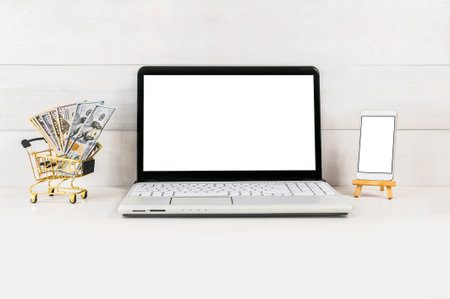 Laptop with blank screen, smartphone on mini easel, and shopping cart filled with US dollars. Concept of online shopping, digital finance, and e-commerce business.の写真素材