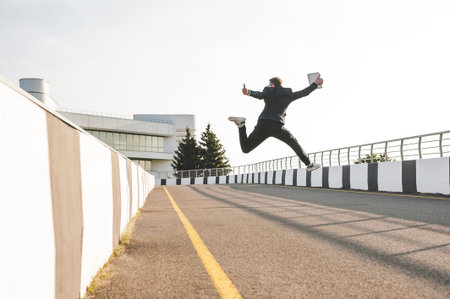 A dynamic shot of a businessman in a suit jumping triumphantly outdoors, holding a tablet and giving...