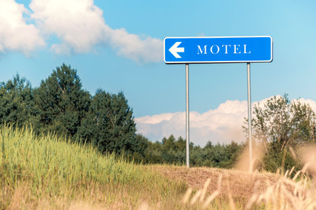 A clear blue motel sign with a directional arrow points left against a backdrop of green trees, a blue sky, and golden grass, perfect for concepts of travel, roadside accommodation, or rural journeys.の写真素材