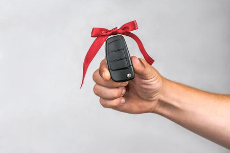 A hand holds a modern black car key adorned with a festive red ribbon against a simple gray...