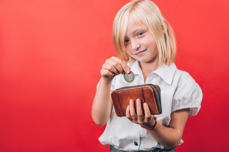 A young girl with blonde hair smiles while placing a coin into a brown wallet, symbolizing children's savings, financial education, and responsible money management.の写真素材