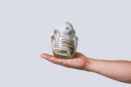 A hand holds a glass jar filled with US dollar banknotes, symbolizing family savings, financial...