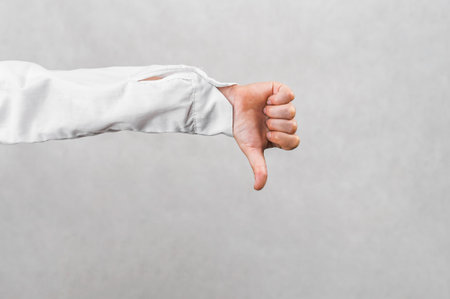 A child's hand in a white shirt sleeve displays a clear thumbs-down gesture against a plain gray background, symbolizing disapproval, disagreement, failure, and negative feedback.の写真素材