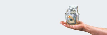 A hand cradle a glass jar filled with US dollar bills, presented on a long banner with ample white...