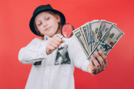 A girl in a black hat examines US dollar banknotes with a magnifying glass. Concept of money, finance, fraud detection, or currency inspection. Red background creates bold visual contrast.の写真素材